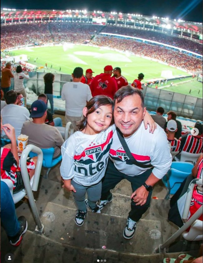 Jura na arquibancada de um estádio lotado, de camisa do São Paulo FC, acompanhado de uma criança
